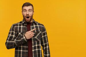 Amazed attractive young bearded man in plaid shirt with opened mouth looks astonished and pointing away to the side over yellow background photo