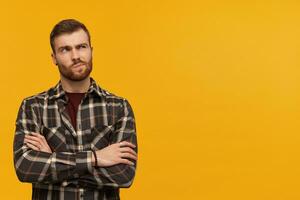 Thoughtful skeptical young man in checkered shirt with beard keeps arms crossed and thinking over yellow background Looking away to the side photo