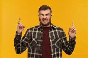 Mad furious young man in plaid shirt with beard pointing up to the sky by both hands and looking at camera over yellow background photo