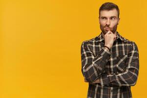 Pensive handsome young bearded man in checkered shirt touching his chin and thinking over yellow background Looking away to the side photo