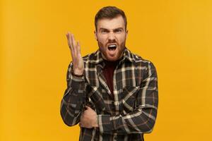 furioso enojado joven hombre en tartán camisa con barba y elevado mano mira agresivo gritos y discutiendo terminado amarillo antecedentes foto