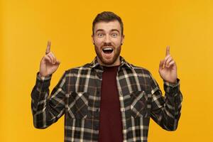 Surprised excited young bearded man in plaid shirt with opened mouth shouting and pointing up to the sky with both hands over yellow background photo