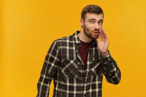 Smiling attractive young man in plaid shirt with beard keeps hand near face and telling a secret isolated over yellow background photo