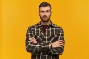 Serious attractive young bearded man in plaid shirt looks confident standing with hands folded and raised brow over yellow background photo