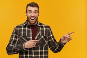 Happy wondered young man in plaid shirt with beard and opened mouth looks amazed and pointing away at empty space by two fingers over yellow background photo
