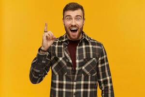 Happy exited young bearded man in plaid shirt shouting pointing up by finger and having an idea isolated over yellow background photo