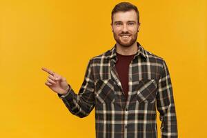 sonriente confidente joven barbado hombre en tartán camisa en pie y señalando lejos a vacío espacio por dedo terminado amarillo antecedentes mirando a cámara foto