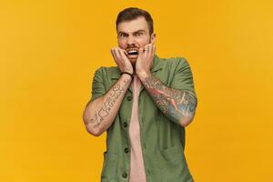 Scared shocked bearded young man with tattoo keeps hands on face and shouting over yellow background photo