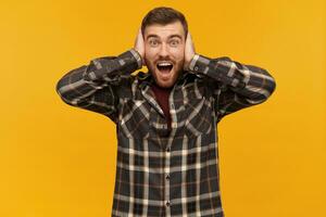 Cheerful excited young man with beard and opened mouth in plaid shirt covered his ears by hands and shouting over yellow background photo