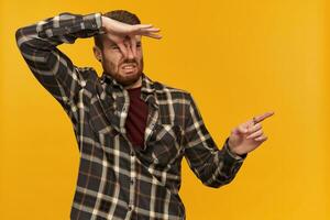 Disgusted unhappy young bearded man in plaid shirt pinches nose by hand feels bad smell or stink and pointing away to the side at empty space over yellow background photo
