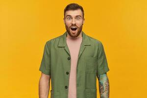 Amazed handsome young man with beard and tattoo standing with opened mouth and looks surprised over yellow background photo