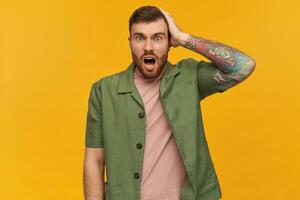 Shocked handsome young man with beard and tattoo keeps hand on head and mouth opened looks emotional and screaming over yellow background photo