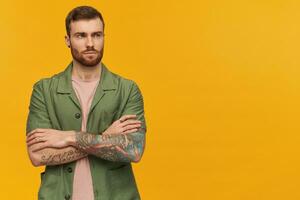 Serious confident young man with beard and tattoo keeps arms crossed and looking away to the side over yellow background photo