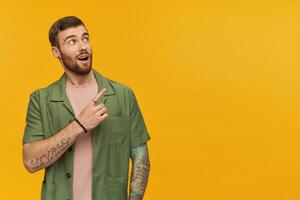 Astonished handsome young man with beard and tattoo looks amazed and pointing away to the side over yellow background photo