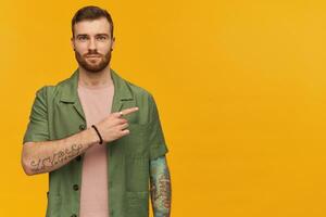 Confident attractive bearded young man with tattoo standing and pointing away to the side at copyspace over yellow background photo