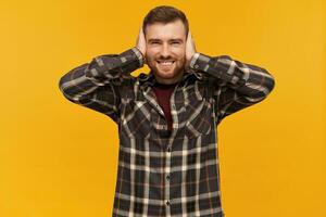 Happy handsome bearded young man in checkered shirt covered his ears by hands and looking at camera over yellow background photo