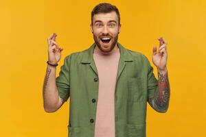 Happy excited young man with beard and tattoo looks inspired keeps fingers crossed and making a wish over yellow background photo