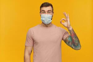 Excited tattooed young man in pink t shirt and virus protective mask on face against corona virus with beard standing and showing ok sign over yellow background photo