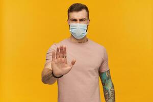 Strong young man in pink t shirt and virus protective mask on face against corona virus with beard and tattoo showing stop gesture by his hand over yellow background photo