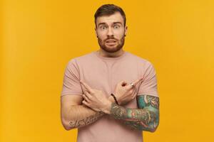 Confused handsome tattooed young bearded man in pink t shirt making x shape with arms and and pointing at both sides by fingers over yellow background photo