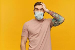 Astonished tattooed young man in pink t shirt and virus protective mask on face against corona virus with beard keeps hand on head and looks shocked over yellow background photo