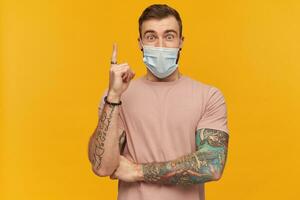 emocionado joven hombre en rosado t camisa y virus protector máscara en cara en contra corona virus con barba y tatuaje en mano señalando arriba y teniendo un idea terminado amarillo antecedentes foto