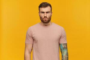 Portrait of angry handsome tattooed young man in pink t shirt with beard looks tensed and irritated over yellow background Standing and looking at camera photo