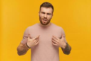enojado confuso tatuado joven barbado hombre en rosado t camisa en pie y señalando a él mismo por ambos manos terminado amarillo antecedentes siente enojado foto
