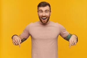 Cheerful attractive young bearded man with tattoo on hand in pink t shirt looks excited and pointing down by fingers of both hands over yellow background photo