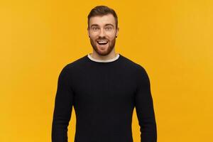 Cheerful amazed young man with beard and opened mouth in black long sleeve feels excited and looks surprised over yellow background photo