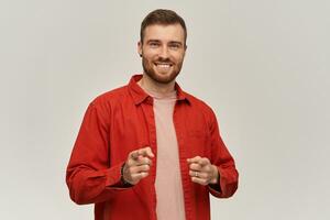 contento atractivo joven barbado hombre en rojo camisa mira confidente sonriente y señalando en usted a cámara terminado blanco antecedentes foto