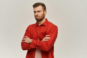 Upset unhappy young man in red shirt with beard feels sad and keeps arms crossed over white background photo