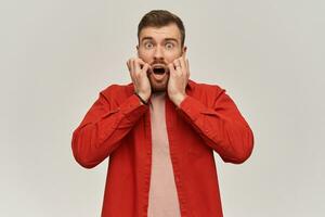 Frightened terrified young bearded man in red shirt looks afraid and screaming over white background photo