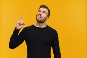 Unhappy displeased young man with beard in black long sleeve looks irritated and pointing up to copy space over yellow background photo