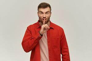 Angry irritated young bearded man in red shirt standing and showing silence sign over white background Looking at camera photo