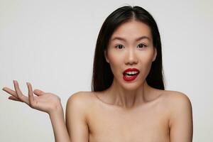 Puzzled young attractive female with festive makeup raising confusedly palm while looking wonderingly at camera, standing over white background with naked shoulders photo