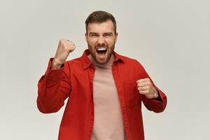 Cheerful excited young man in red shirt with beard keeps fists clenched and raised hands and screaming over white background Celebrating victory concept photo