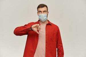 Sad bored young man with beard in red shirt and hygienic mask to prevent infection looking up and showing thumbs down over white background photo