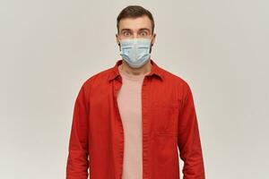 Worried shocked young bearded man in red shirt and virus protective mask on face against corona virus standing and looking at camera over white background photo