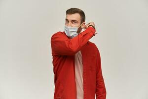 Handsome young man with beard in red shirt trying to take off or put on hygienic mask for preventing infection over white background photo