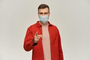 Skeptical young man with beard in red shirt and hygienic mask to prevent infection pointing on camera or you by finger over white background photo