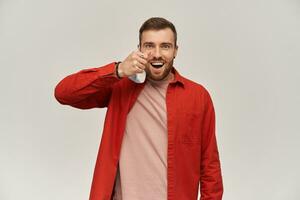 Cheerful young bearded man in red shirt standing and taking off virus protective mask against corona virus from his face over white background photo