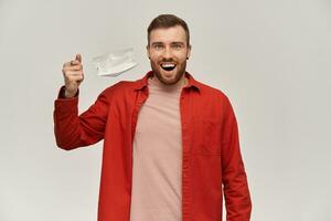 Happy young man with beard in red shirt taking off hygienic mask for preventing infection and breathe deeper over white background photo