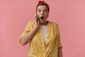 Portrait european charming woman 20s with natural makeup wearing white t-shirt and yellow shirt and red bandana with arms gesticulates on the phone emotion wow surprised looking at you over pink wall photo