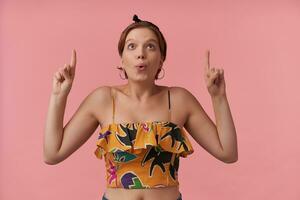 Image of over attractive young woman brown eyed pink background amazing wearing summer crop top slim strap and bandana surprised emotion with arms pointing fingers up emotion wow looking aside up photo