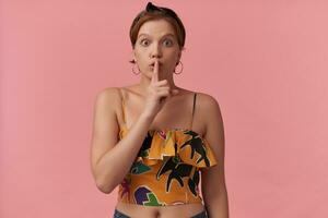 Image of european brown haired youth woman wearing summer outfit ear rings and black bandana arms touching face finger over lips isolated posing over pink background emotion quite secret keep silence photo