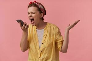 Image european brown haired youth woman in trendy summer outfit and red bandana with arms gesticulates with smart phone emotion shouting out loud scream mad on gadget isolated against pink background photo