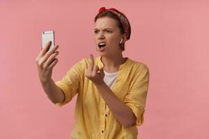 Image european brown haired youth woman white t-shirt and yellow shirt and red bandana posing on self shot emotion rude mad angry unhappy middle finger looking at phone isolated over pink background photo