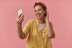 Portrait of attractive young brown eyed wearing yellow shirt and red bandana with arms gesticulates on the phone smiling aside emotion flirting smile happy glad isolated posing over pink background photo