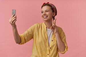 Image of european charming woman 20s with natural makeup wearing trendy summer posing over pink background emotion happy glad smiling making self shot two fingers up arms gesticulates on the phone photo